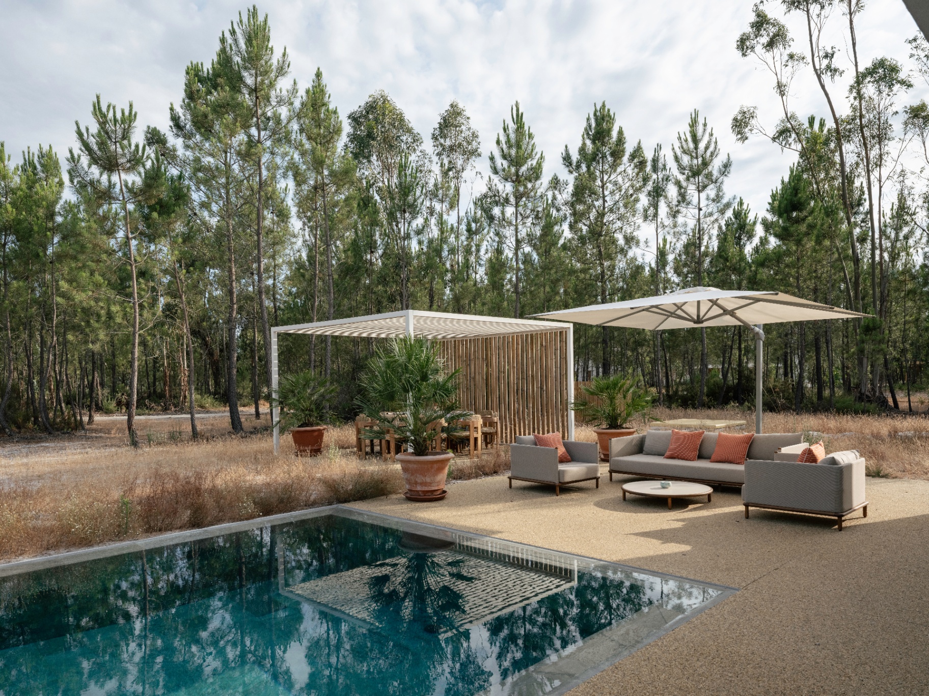 Poolside terrace with lounge seating, pergola, and umbrella surrounded by pine forest.